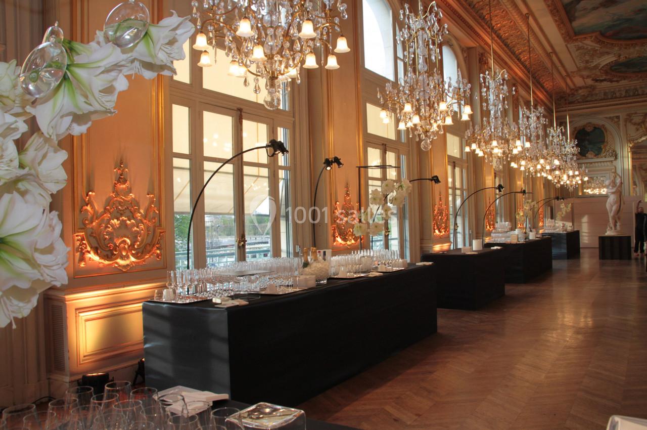 Salle élégante avec lustres, grandes fenêtres et tables noires dressées pour un événement.