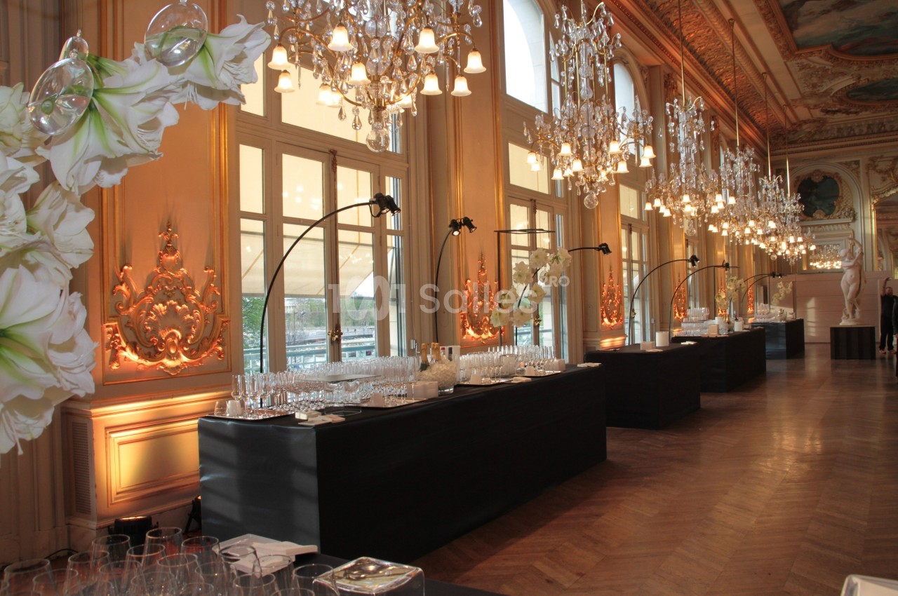 Salle élégante avec lustres, grandes fenêtres et tables noires dressées pour un événement.