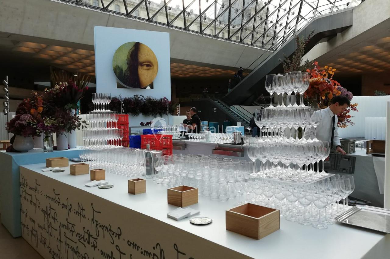 Table dressée avec des verres empilés en pyramide, décor floral et personnel en arrière-plan dans un espace moderne.