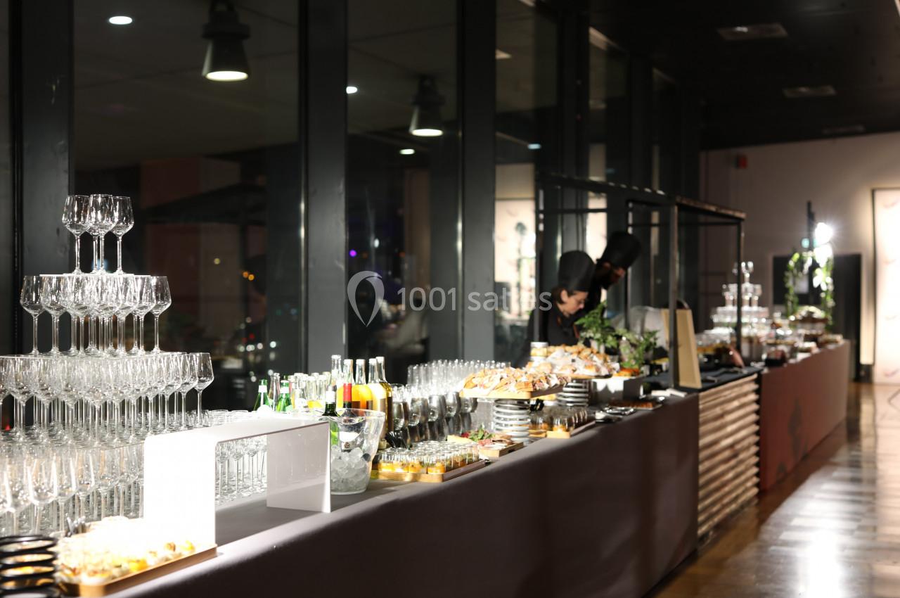 Buffet élégant avec verres empilés, boissons et plats variés dans une salle lumineuse aux grandes baies vitrées.