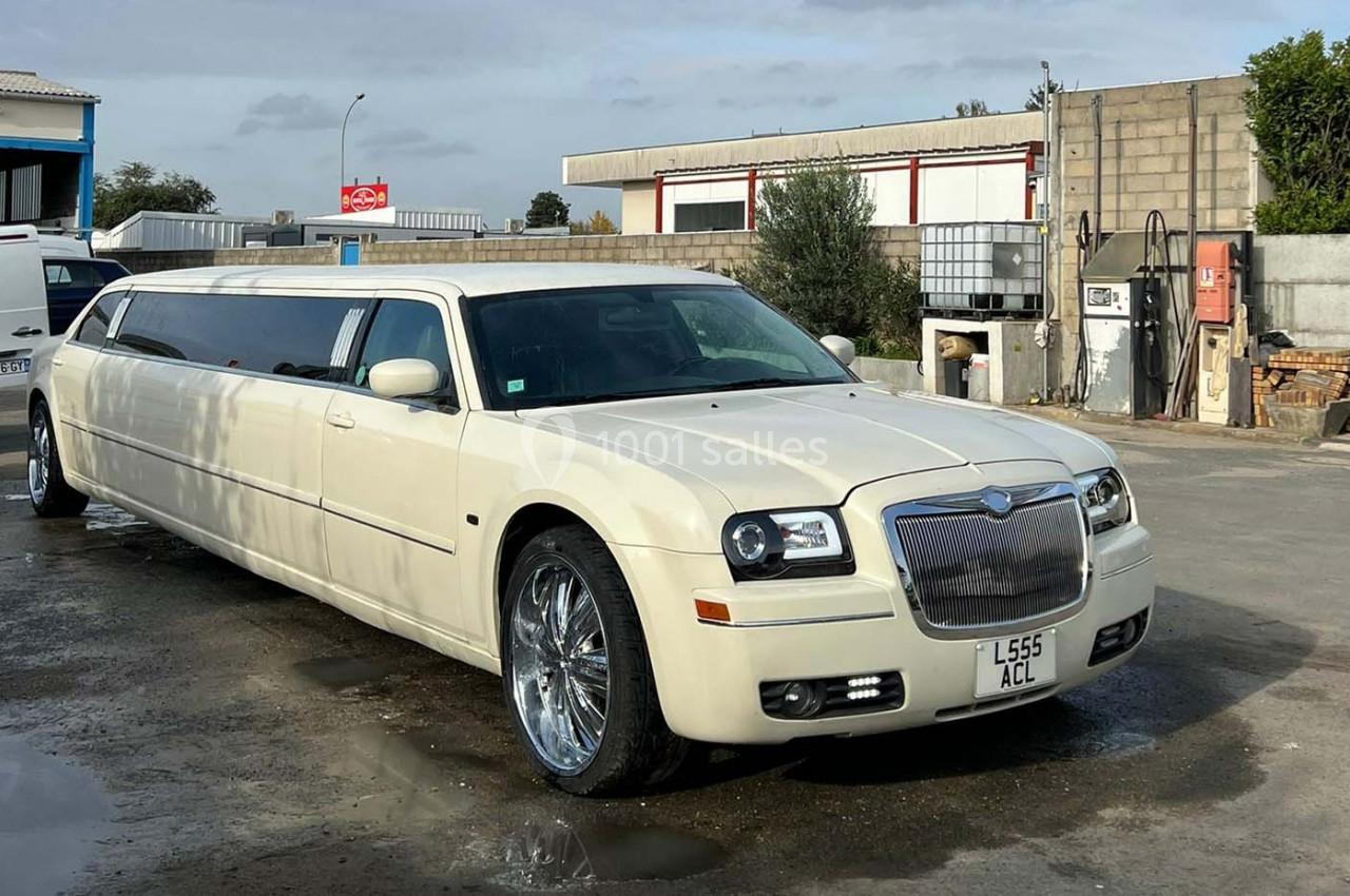 Limousine blanche stationnée sur un sol bétonné, entourée de bâtiments industriels et d'équipements extérieurs.