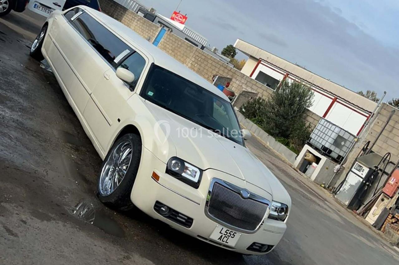 Limousine blanche garée sur un sol humide devant des bâtiments industriels en arrière-plan.