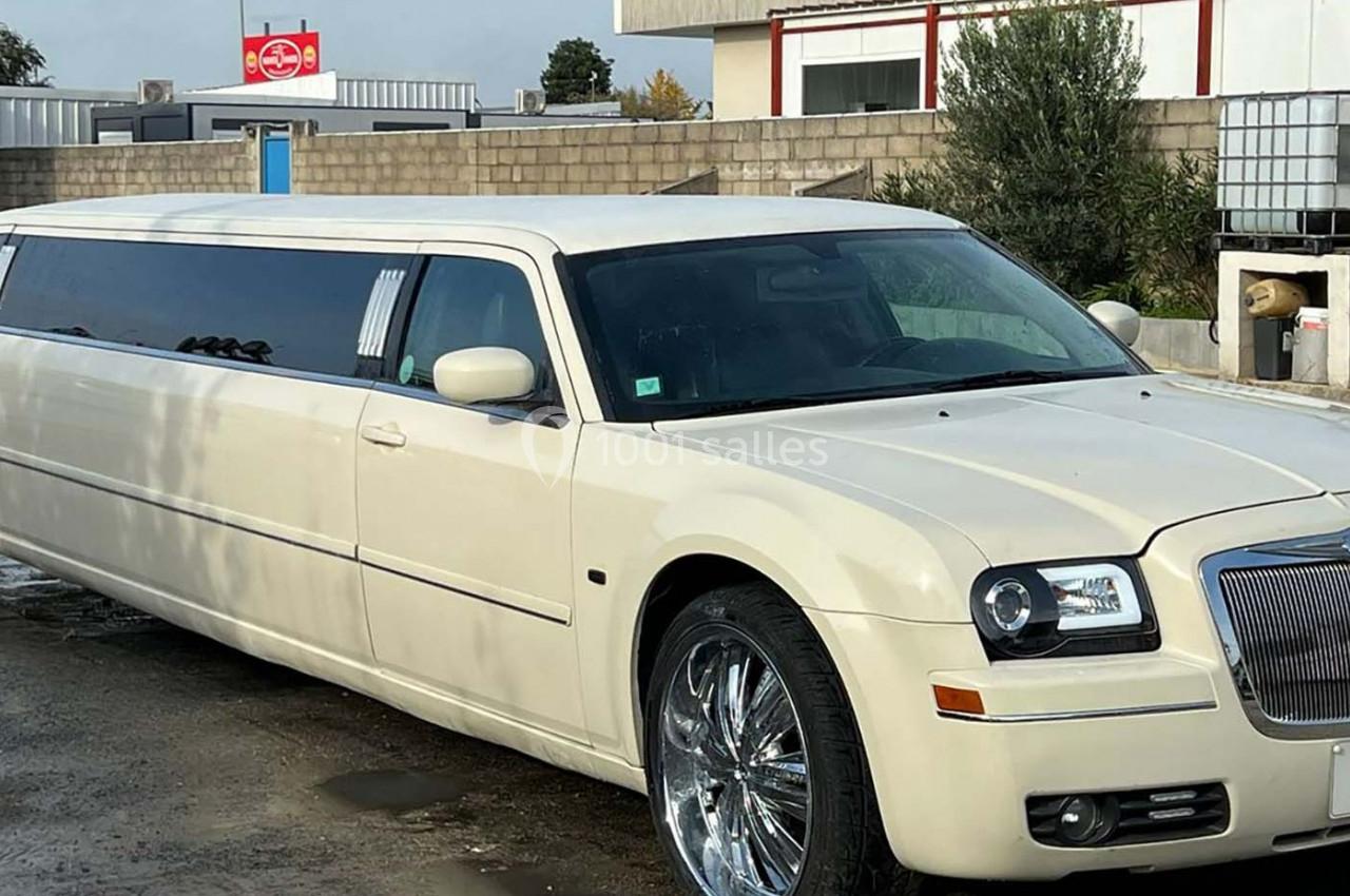 Limousine blanche stationnée sur un sol humide devant un bâtiment et un panneau publicitaire en arrière-plan.