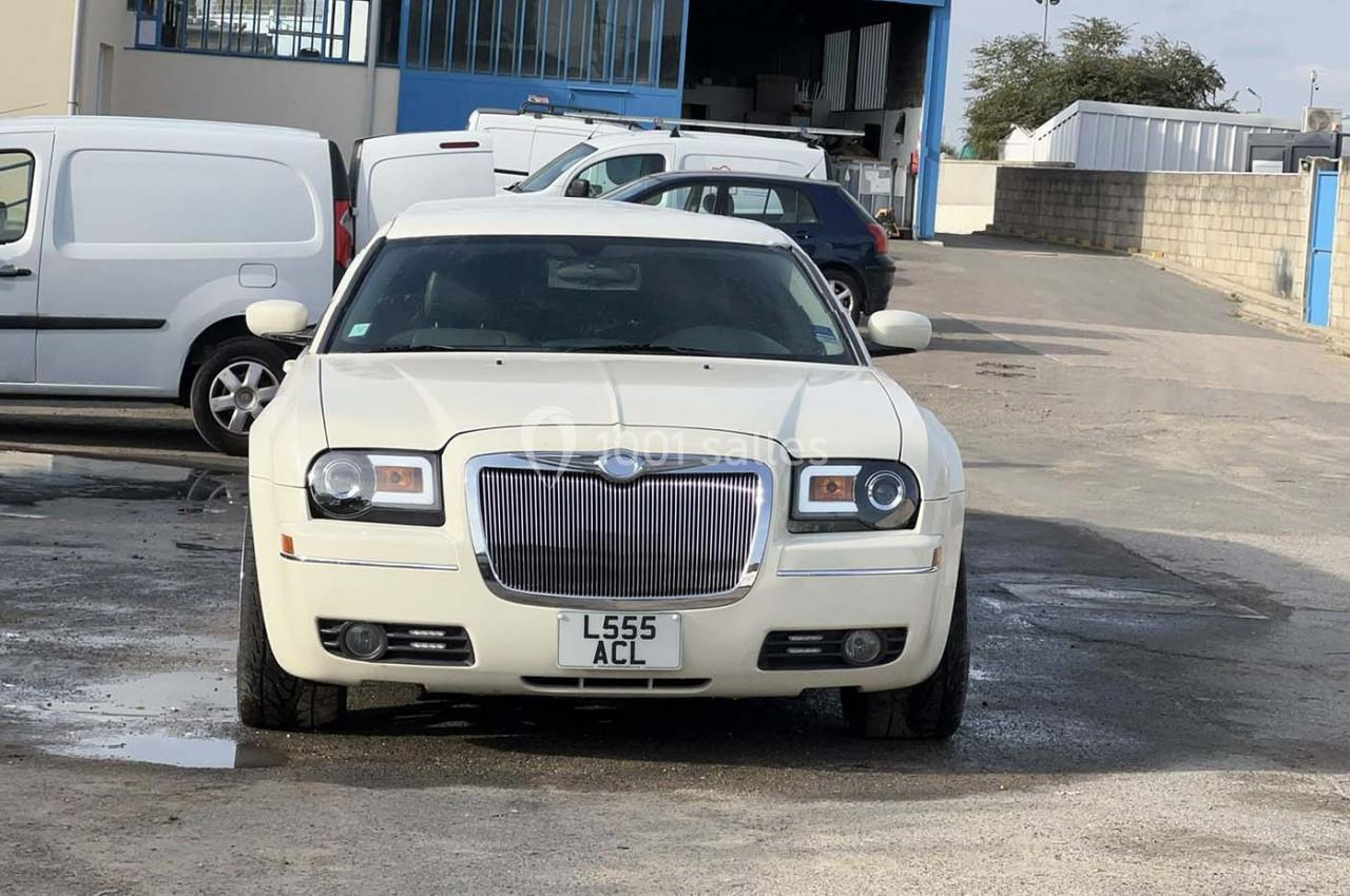 Voiture blanche garée sur un parking devant un bâtiment industriel, entourée de quelques véhicules utilitaires.