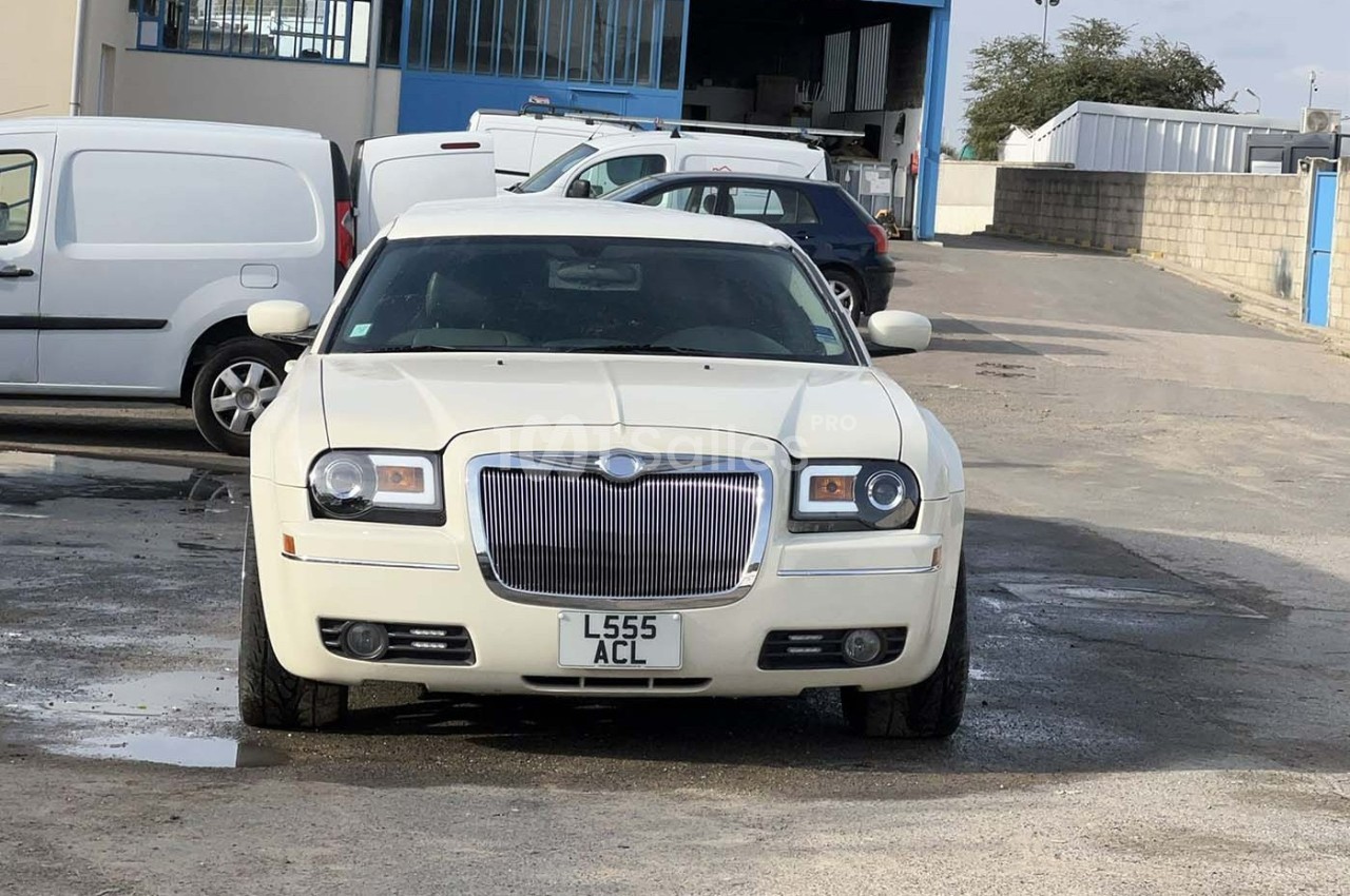 Voiture blanche garée sur un parking devant un bâtiment industriel, entourée de quelques véhicules utilitaires.