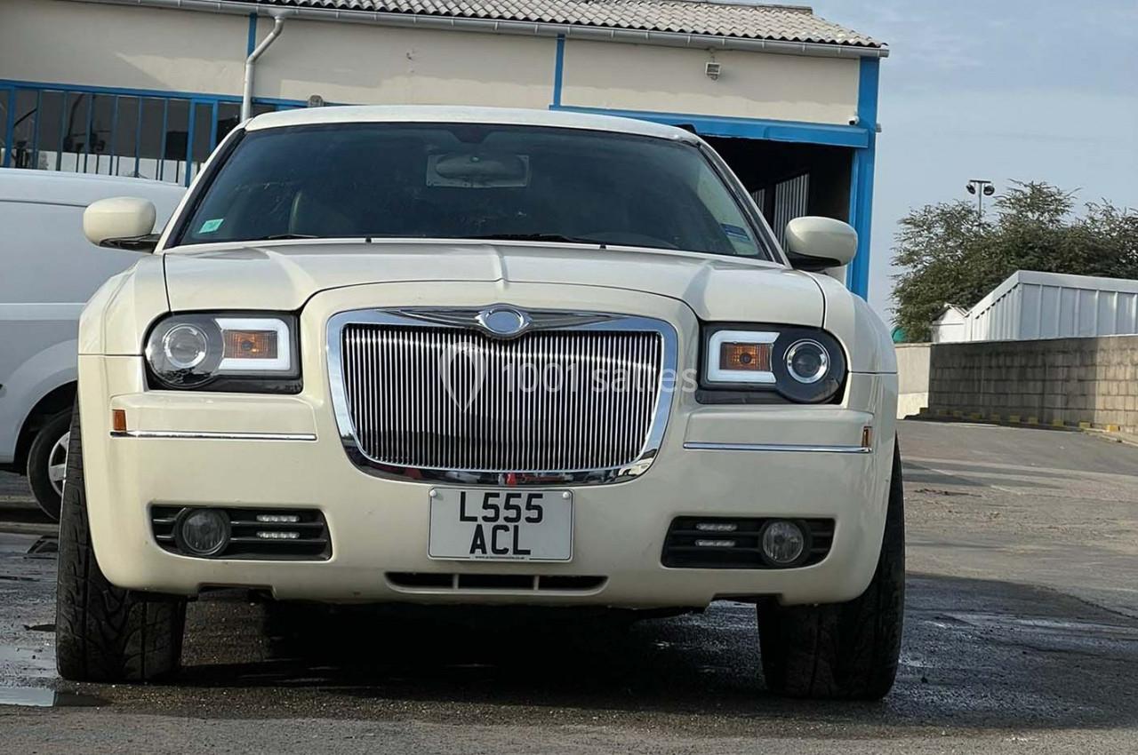 Voiture blanche garée sur un parking, vue de face, avec un bâtiment industriel en arrière-plan.