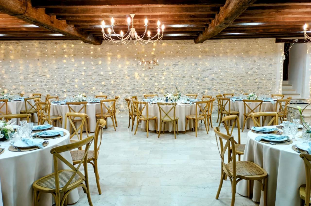 Salle de réception décorée avec des tables rondes dressées, chaises en bois et guirlandes lumineuses sur un mur en pierre.