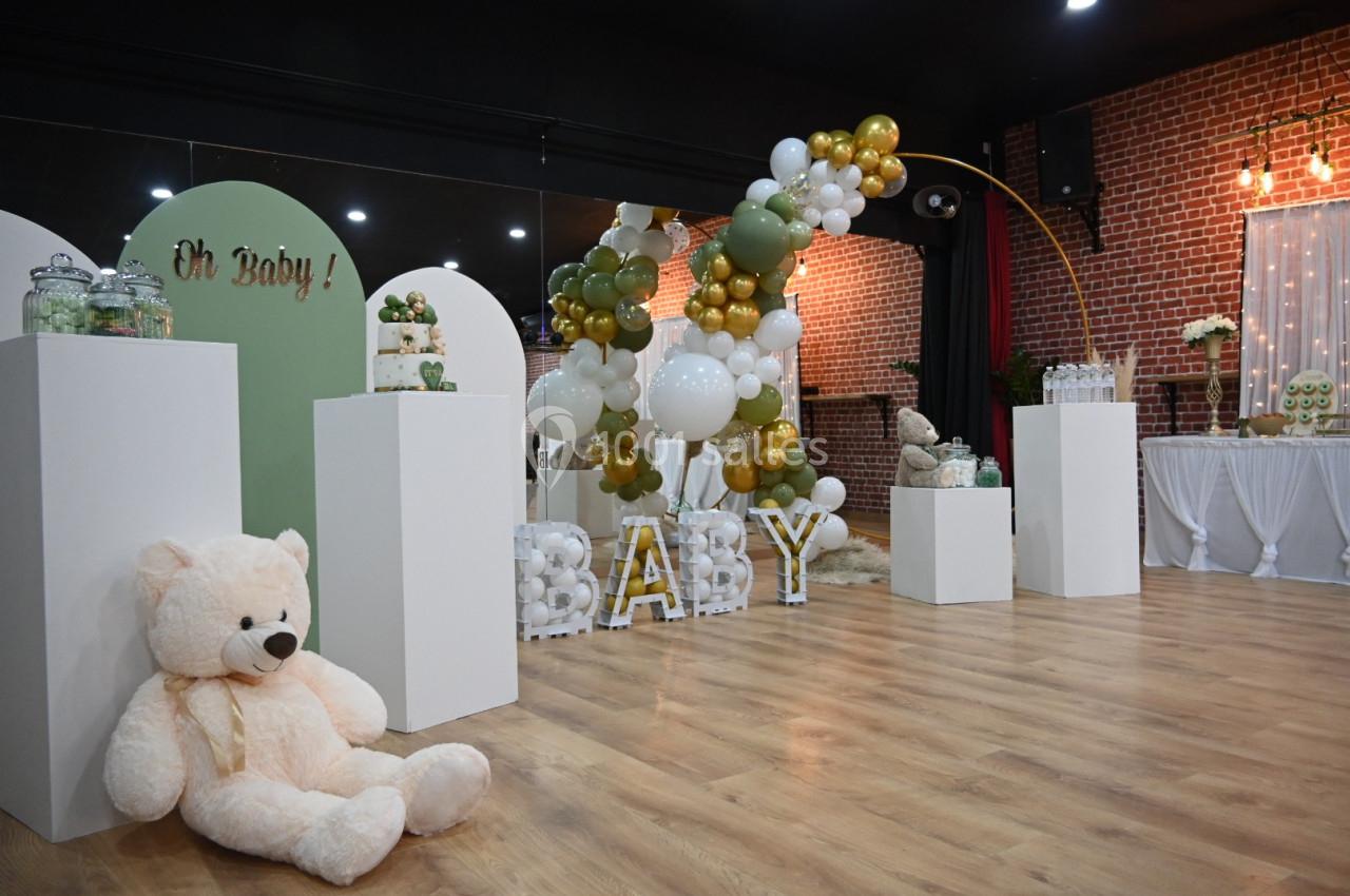 Décoration de fête avec ballons blancs et dorés, lettres ’BABY’, ours en peluche et table garnie dans une salle en briques.