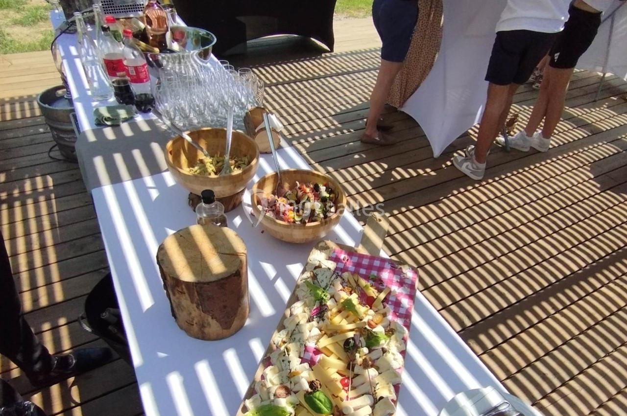Buffet en extérieur avec planches de fromage, salades et boissons disposées sur une table blanche ensoleillée.