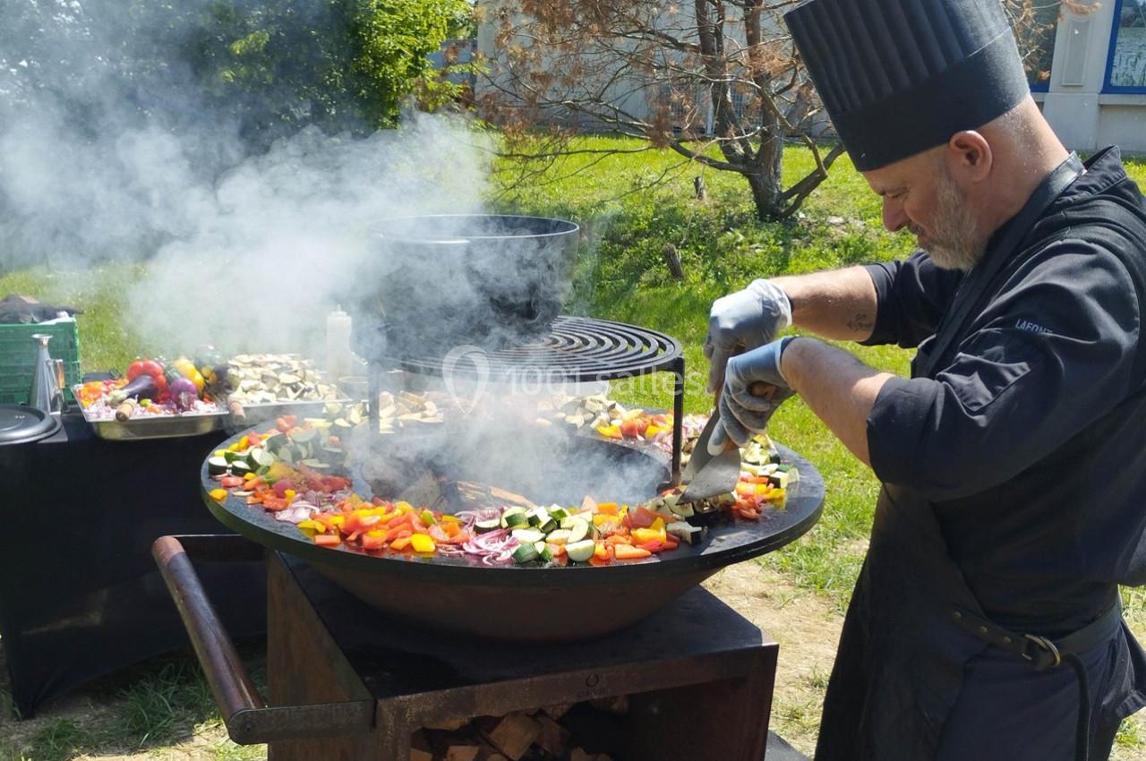 Un chef cuisine des légumes colorés sur une grande plancha ronde en extérieur, entouré de fumée.
