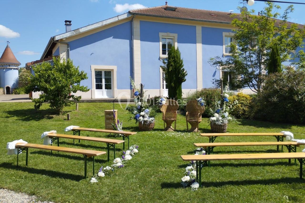 Disposition d'une cérémonie en plein air avec bancs en bois, décor floral et une maison bleue en arrière-plan.