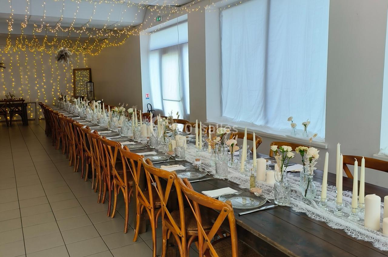 Salle décorée pour un événement avec une longue table en bois, chaises assorties et guirlandes lumineuses suspendues.