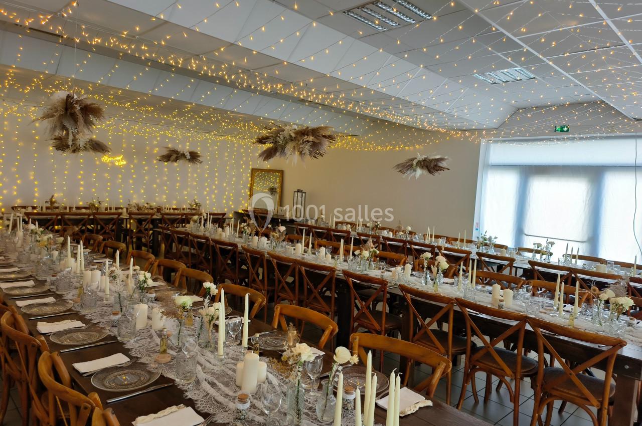 Salle de réception décorée avec des guirlandes lumineuses, tables alignées avec nappes en dentelle et bougies.
