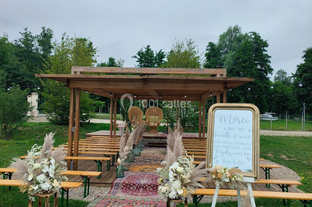 Cérémonie en extérieur avec arche en bois, tapis colorés, bancs en bois et décorations florales.