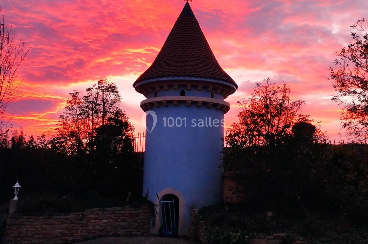 Tour bleue avec un toit en tuiles rouges, entourée de végétation, sous un ciel rouge vif au coucher du soleil.