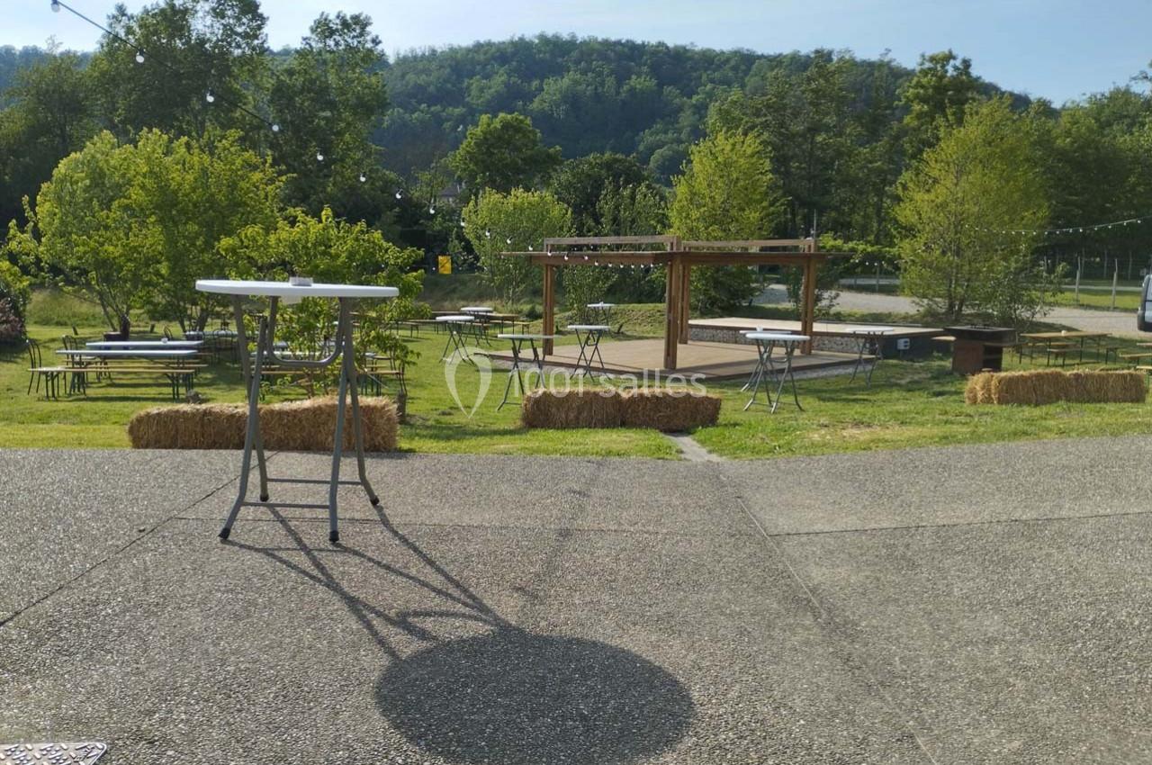 Espace extérieur avec tables hautes, bottes de paille et scène en bois, entouré de verdure et collines.