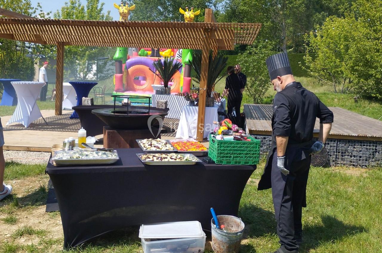 Un chef en tenue noire prépare un buffet en plein air avec des plats disposés sur une table devant un chapiteau coloré.