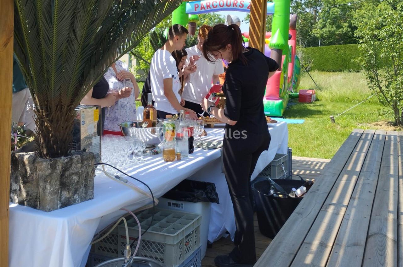 Une femme prépare des boissons sur une table dressée en extérieur lors d'un événement avec des invités et un château…