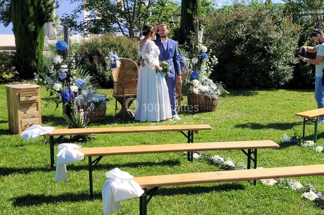 Un couple de mariés debout dans un jardin décoré, entouré de fleurs blanches et bleues, avec des bancs en bois au premier…