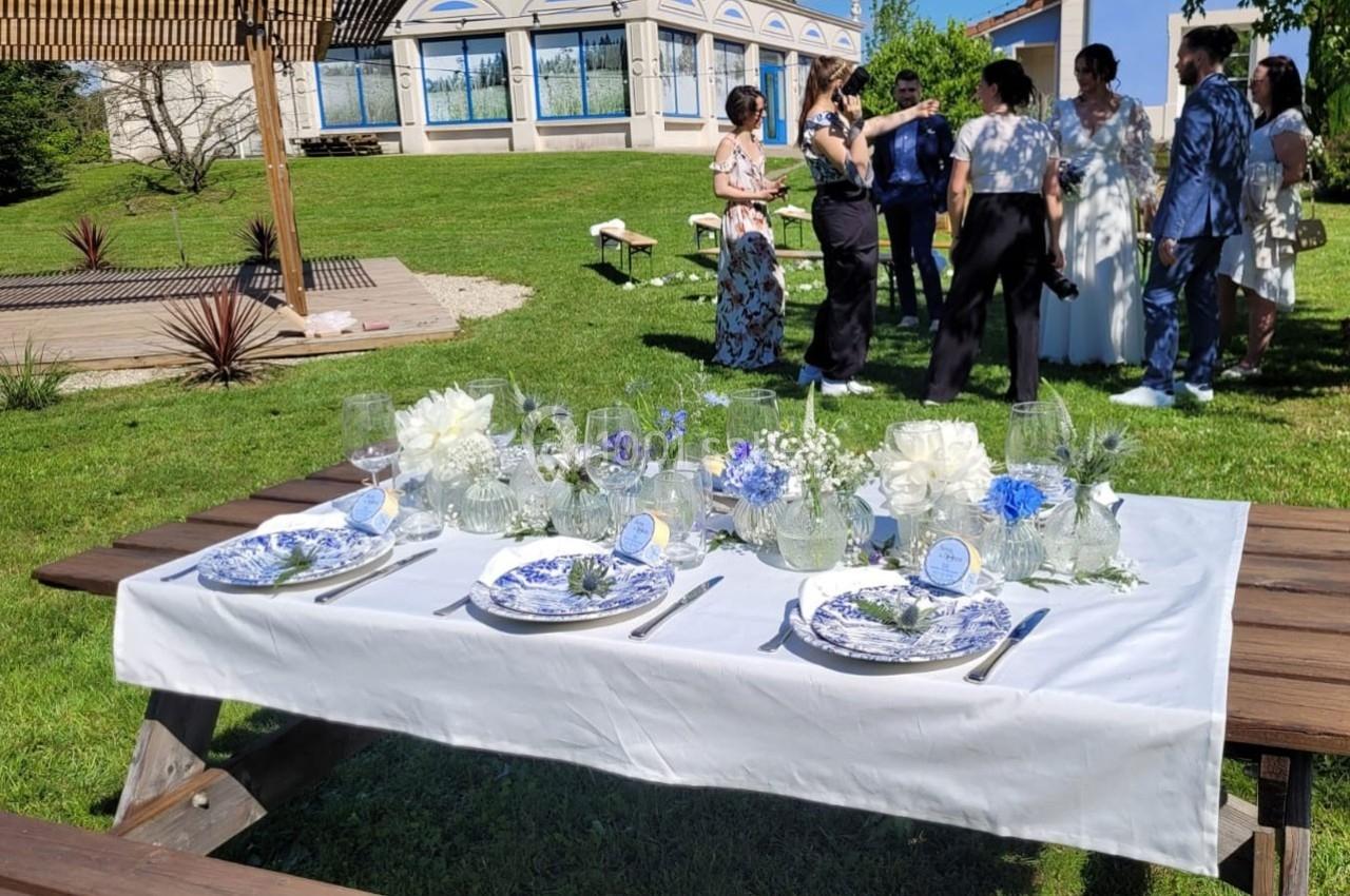 Table de réception extérieure décorée de fleurs blanches et bleues, avec des invités discutant en arrière-plan.