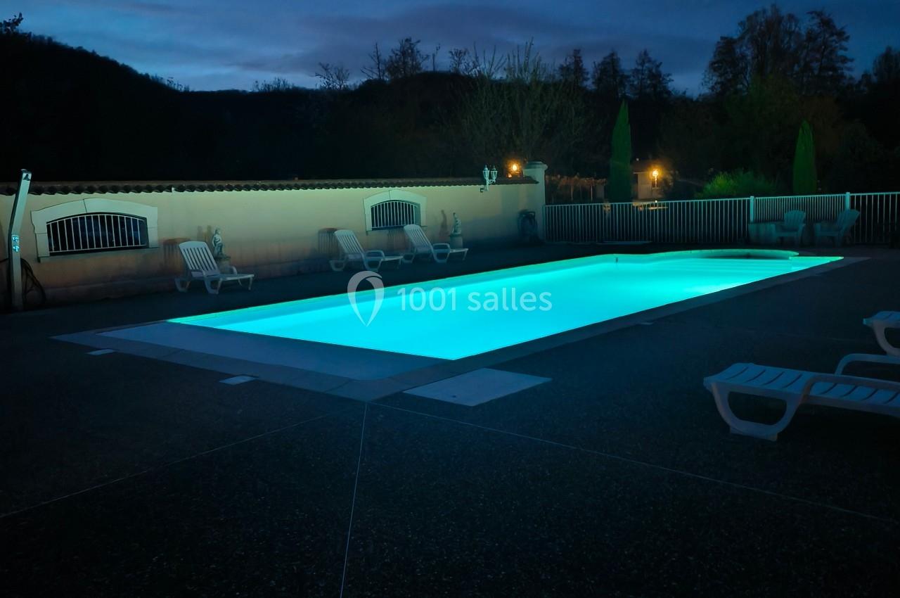 Piscine extérieure éclairée de nuit, entourée de chaises longues et d'arbres en arrière-plan.