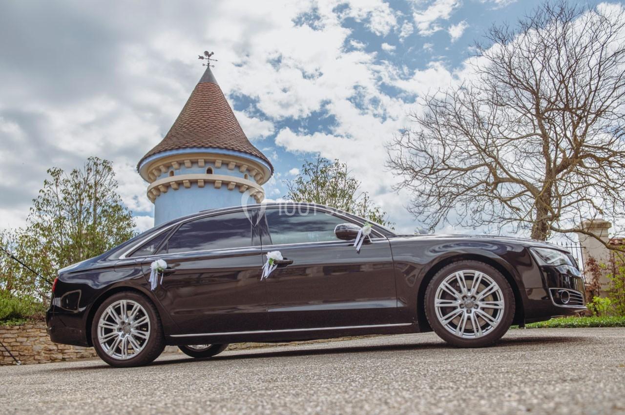 Voiture noire de luxe stationnée devant une tour en pierre avec un toit conique, sous un ciel partiellement nuageux.