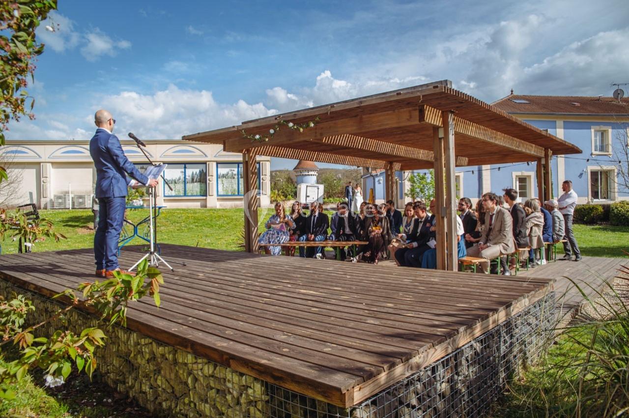 Un homme en costume parle à un groupe de personnes assises sous une pergola en bois dans un espace extérieur.