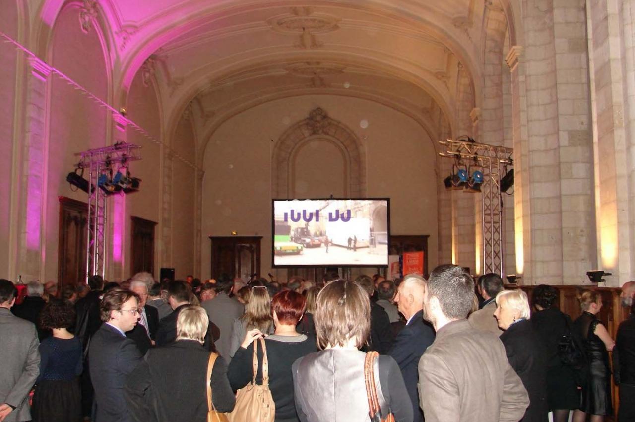Des personnes assistent à un événement dans une grande salle avec un écran géant et un éclairage coloré.