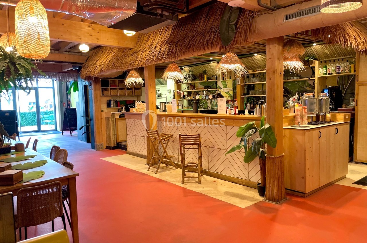 Intérieur d'un café tropical avec comptoir en bois, tabourets, plantes et éclairage chaleureux sous un toit en chaume.
