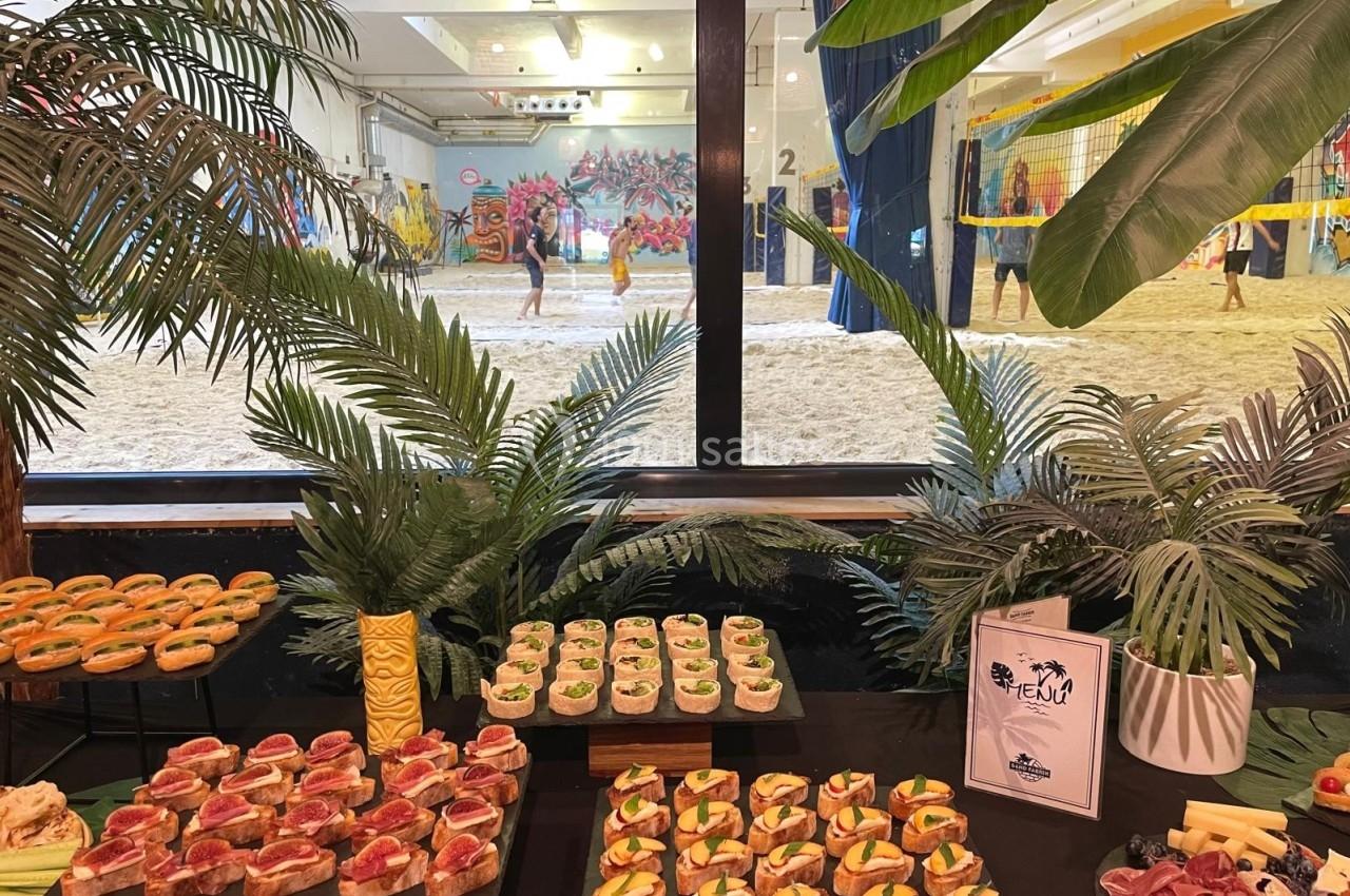 Buffet de canapés variés disposés devant une baie vitrée donnant sur un terrain de beach-volley en intérieur.