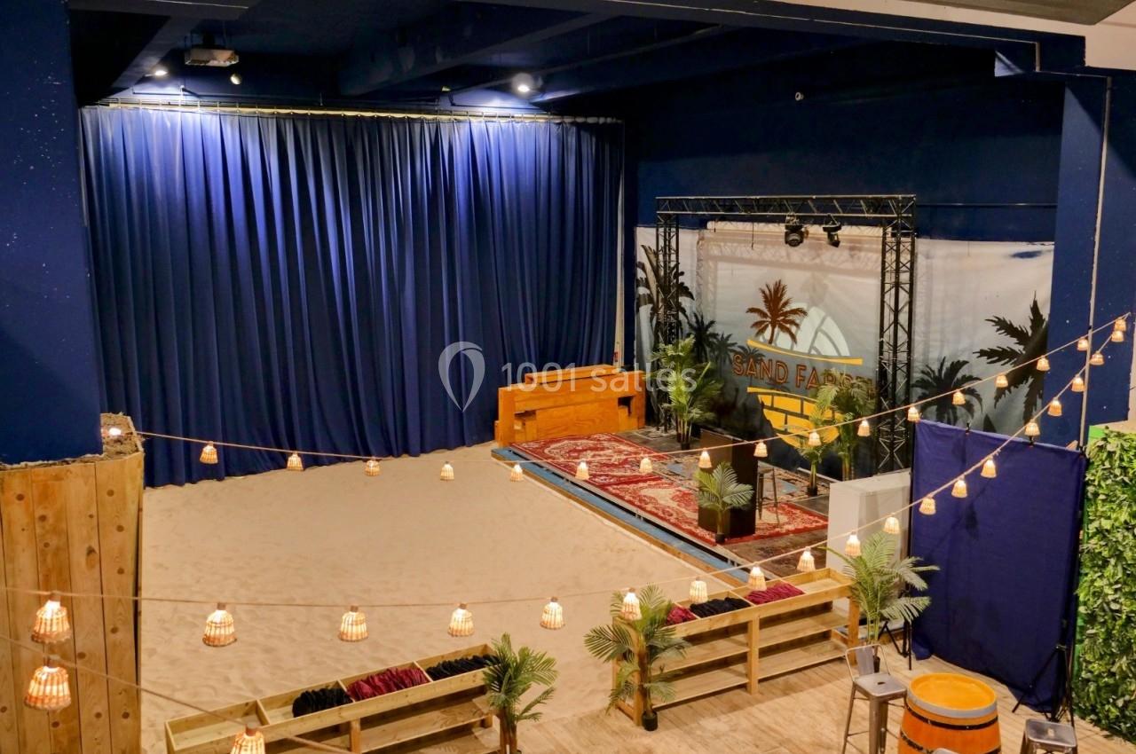 Salle intérieure décorée avec un sol en sable, des guirlandes lumineuses, des bancs en bois et une scène avec rideaux bleus.