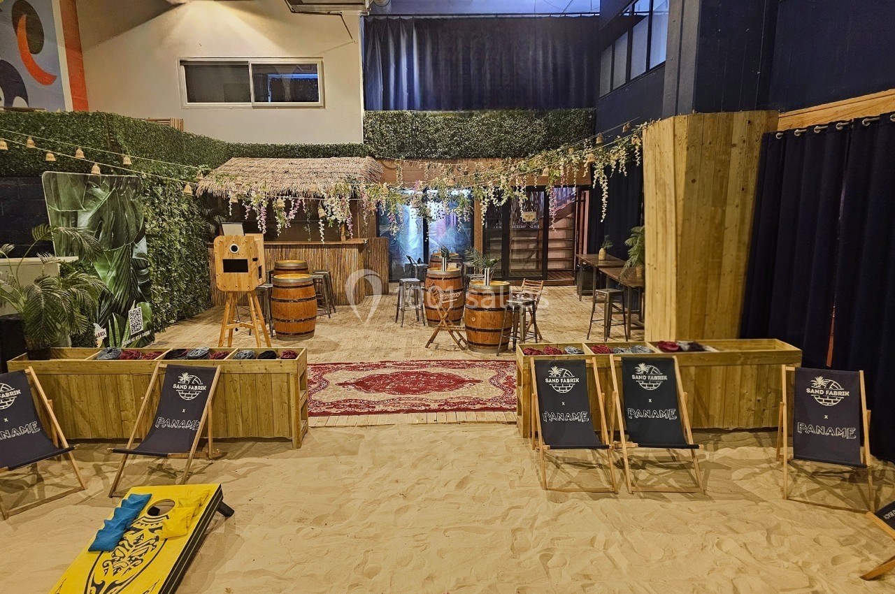 Espace intérieur aménagé avec du sable, des tables en tonneaux, des plantes et des jeux, ambiance conviviale et détendue.