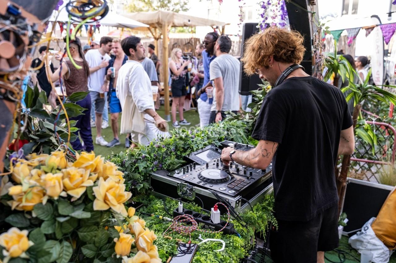 Un DJ mixe en plein air lors d'un événement festif, entouré de végétation et de participants discutant.