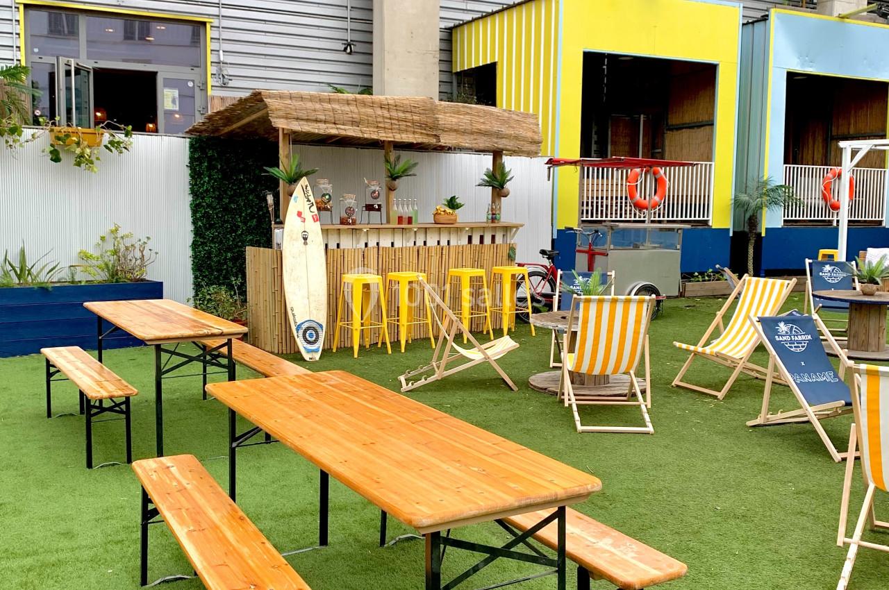 Tables en bois, chaises longues et bar en extérieur sur une terrasse avec gazon artificiel devant un bâtiment.