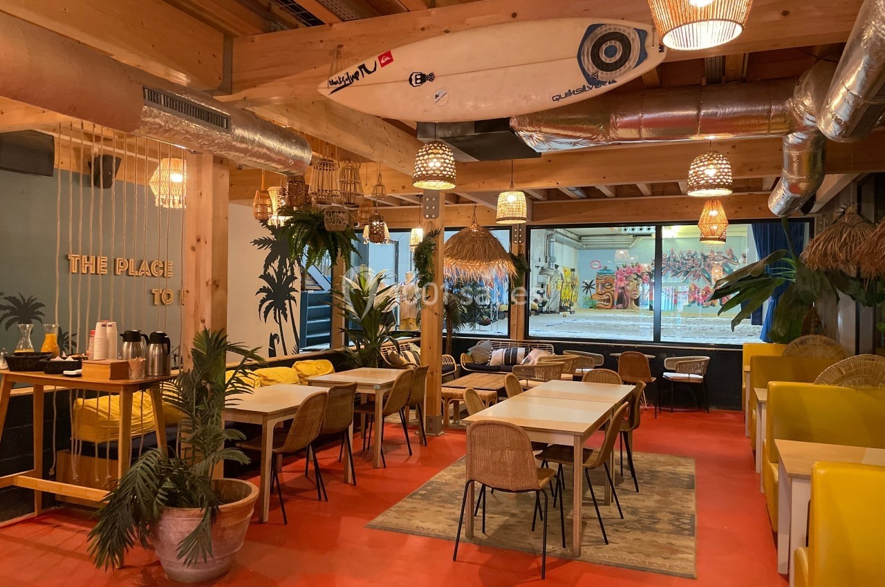 Salle lumineuse avec tables en bois, chaises en rotin, décor tropical et planche de surf suspendue au plafond.