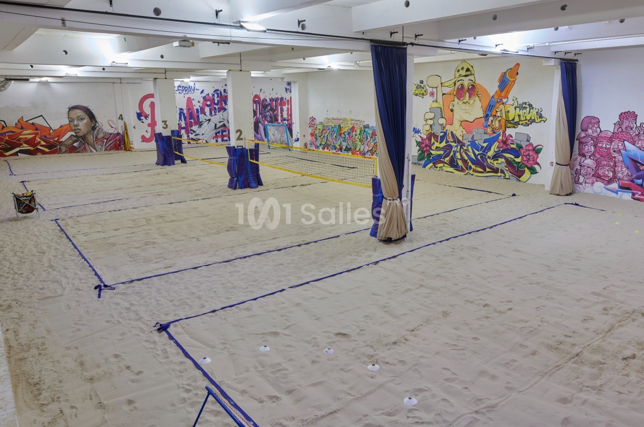 Terrain de beach-volley intérieur avec sol en sable, séparé par des filets et entouré de murs décorés de graffitis colorés.