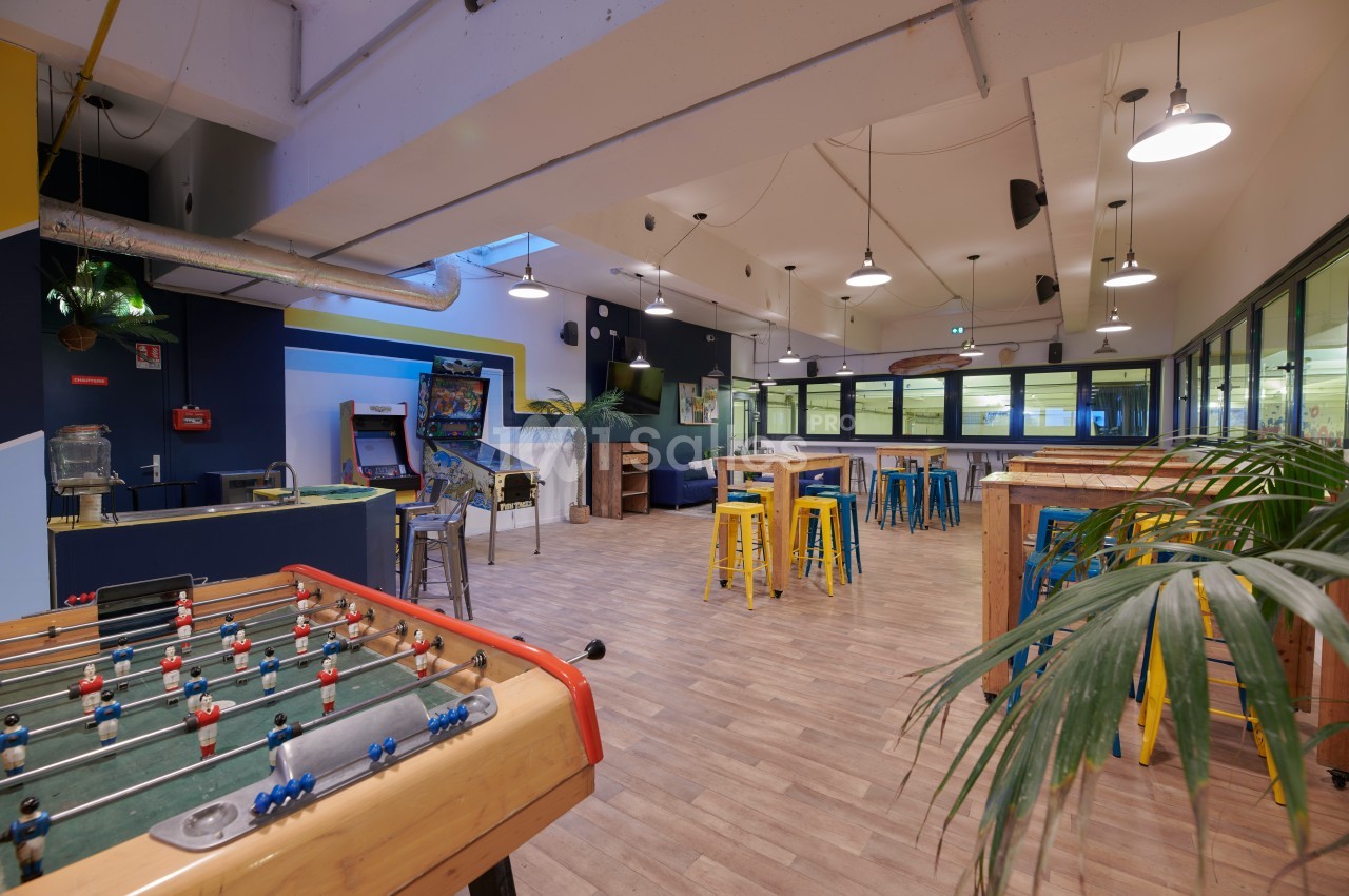 Salle de détente avec baby-foot, tables hautes, tabourets colorés et jeux d'arcade dans un espace lumineux.