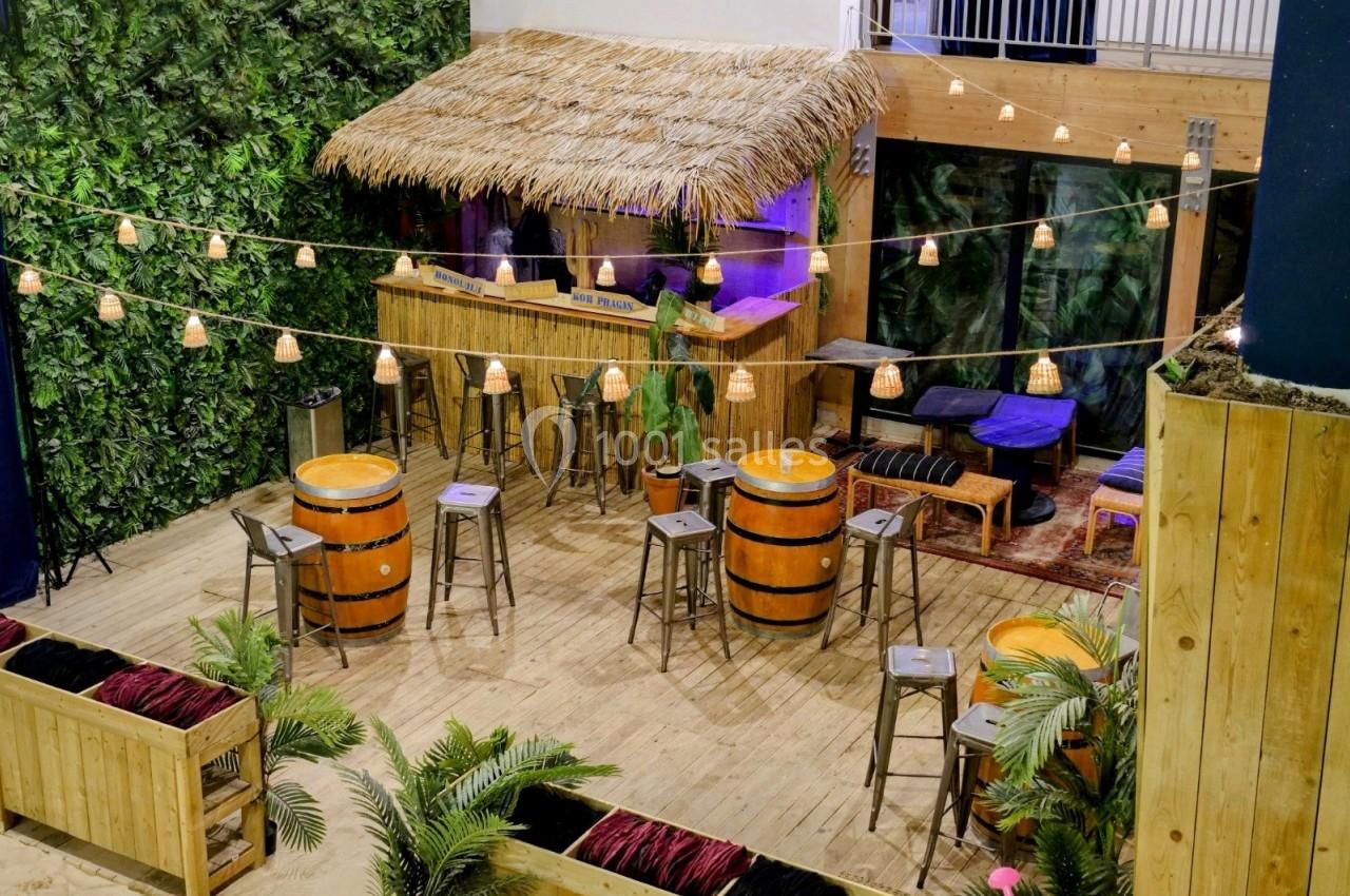 Espace aménagé avec un bar en toit de chaume, des tonneaux, des tabourets et des guirlandes lumineuses, ambiance tropicale.