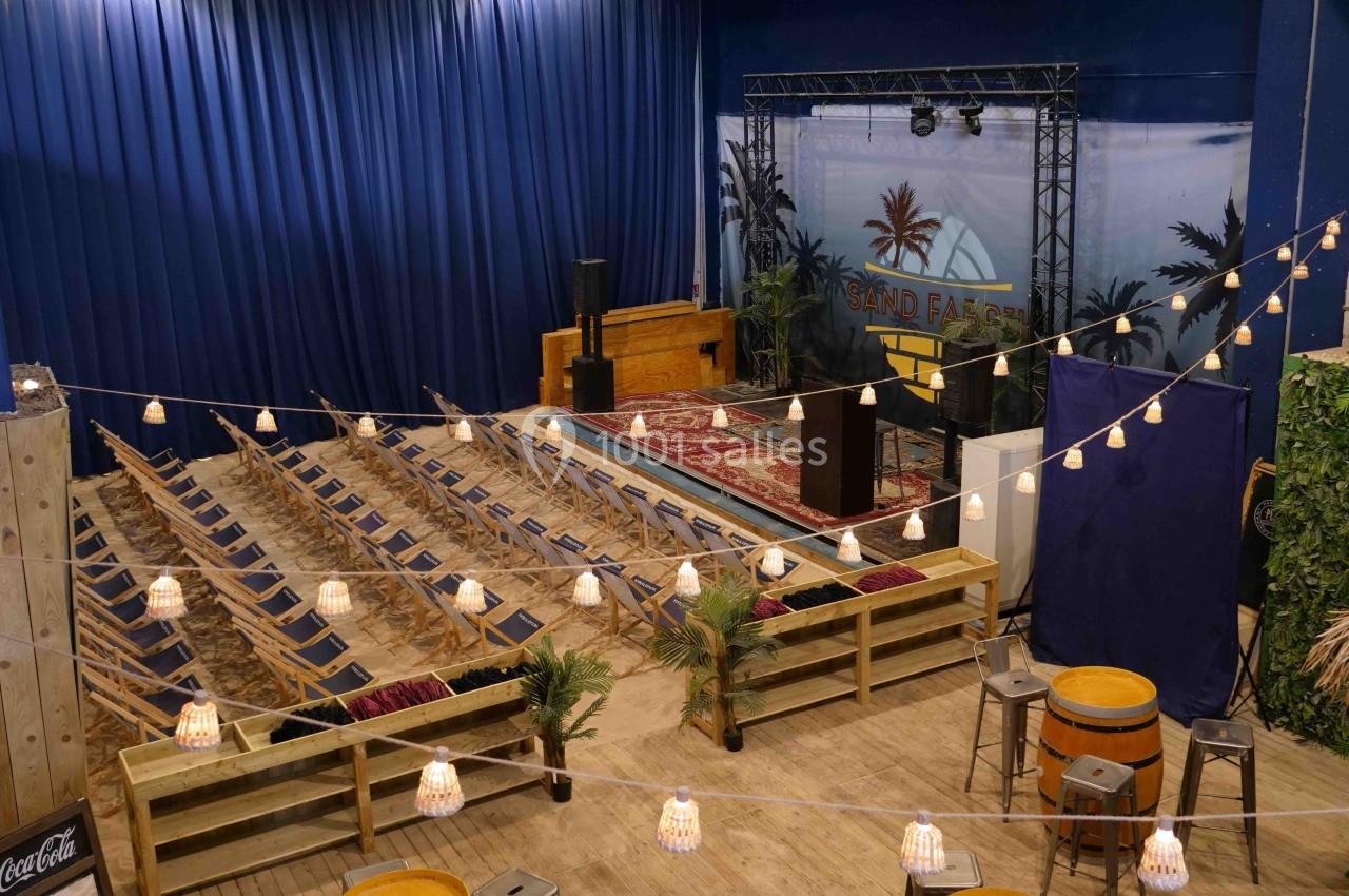 Salle aménagée avec des chaises pliantes, une scène décorée et des guirlandes lumineuses suspendues.