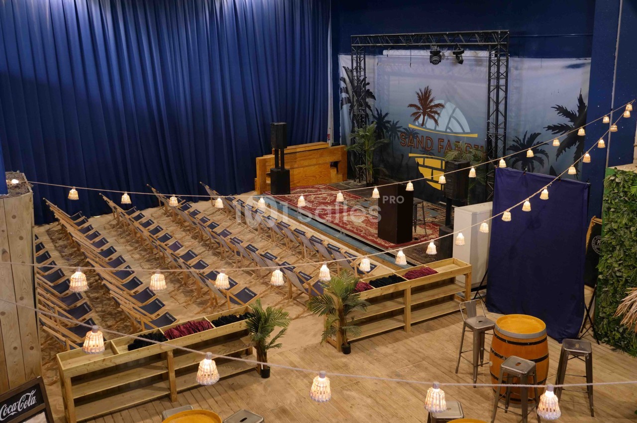 Salle aménagée avec des chaises pliantes, une scène décorée et des guirlandes lumineuses suspendues.
