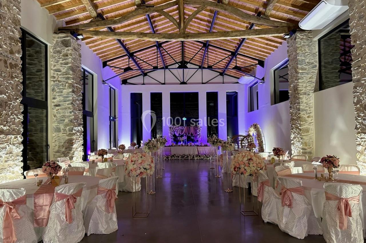 Salle de réception décorée avec des tables rondes ornées de nappes blanches et roses, éclairage chaleureux et arc floral.