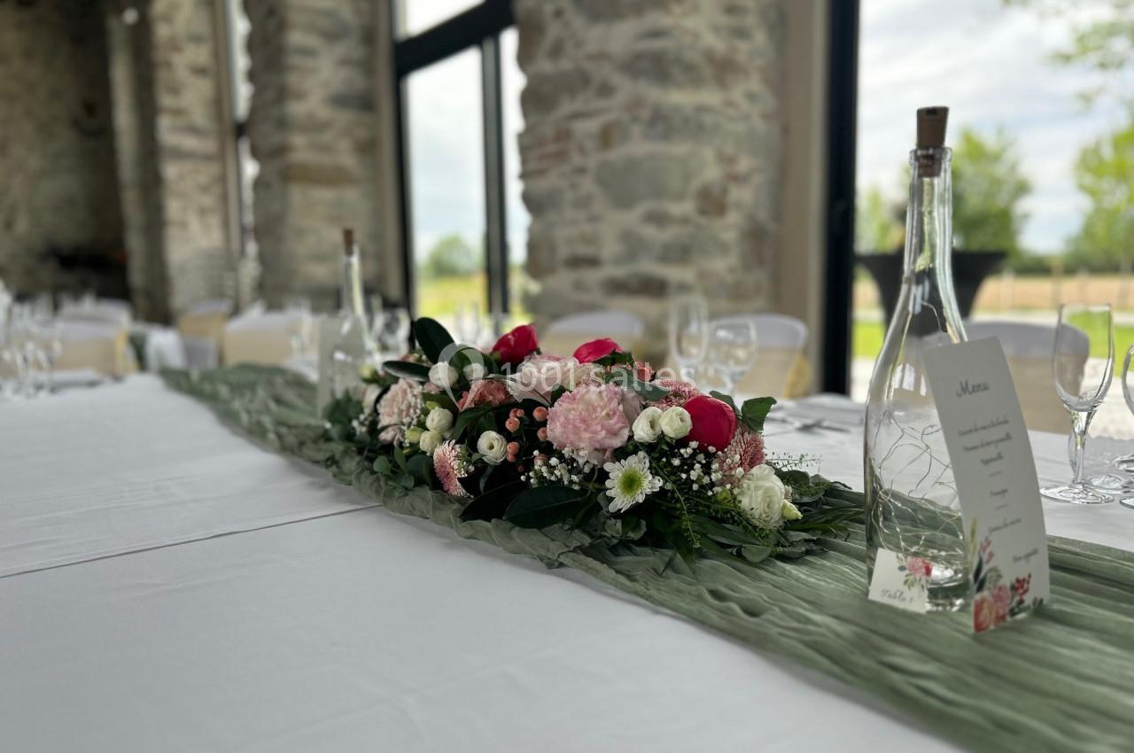 Centre de table floral avec roses et feuillage, posé sur un chemin de table vert dans une salle lumineuse en pierre.