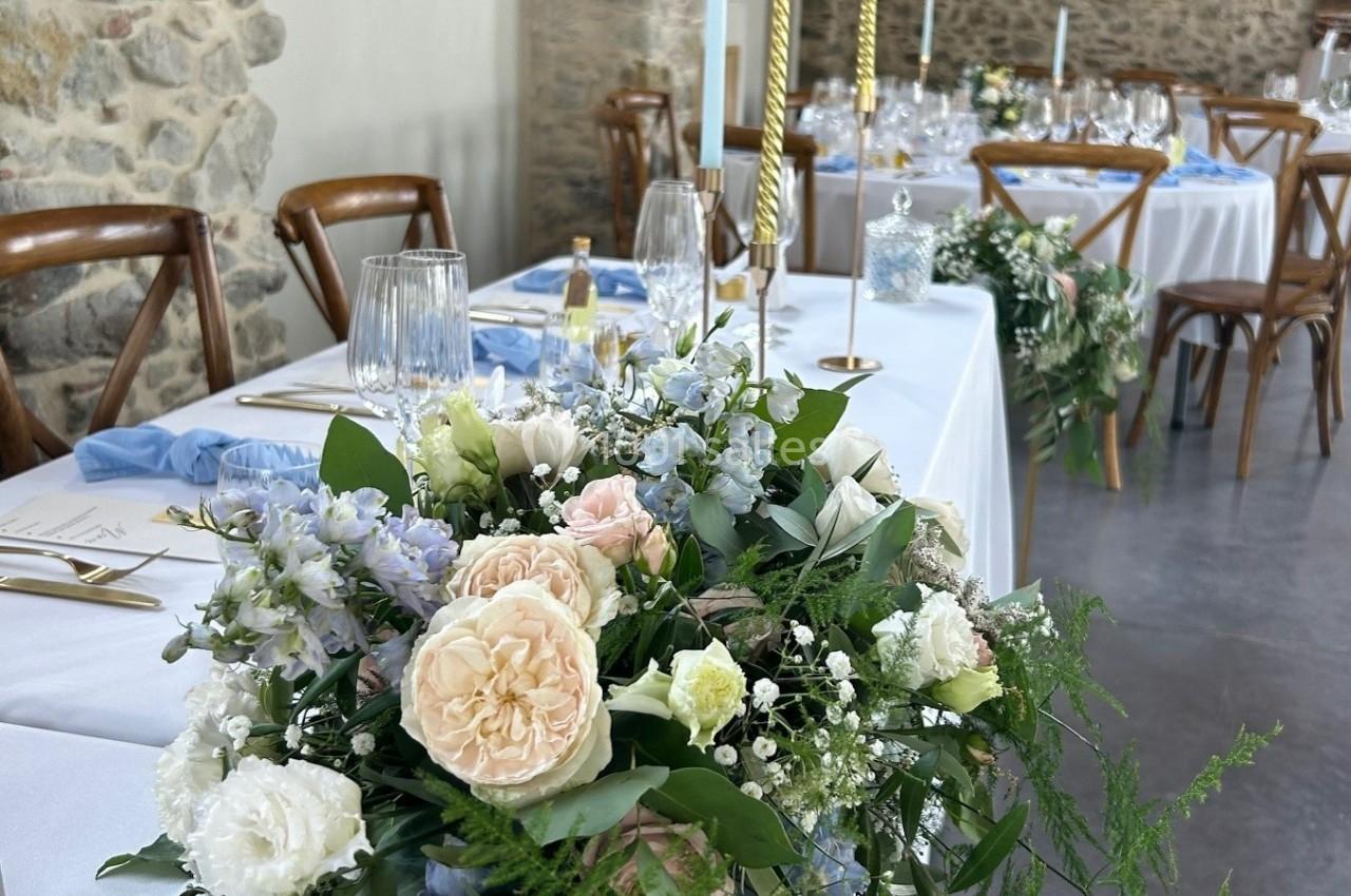 Décoration florale avec roses, gypsophiles et feuillage sur une table de réception dans une salle en pierre.