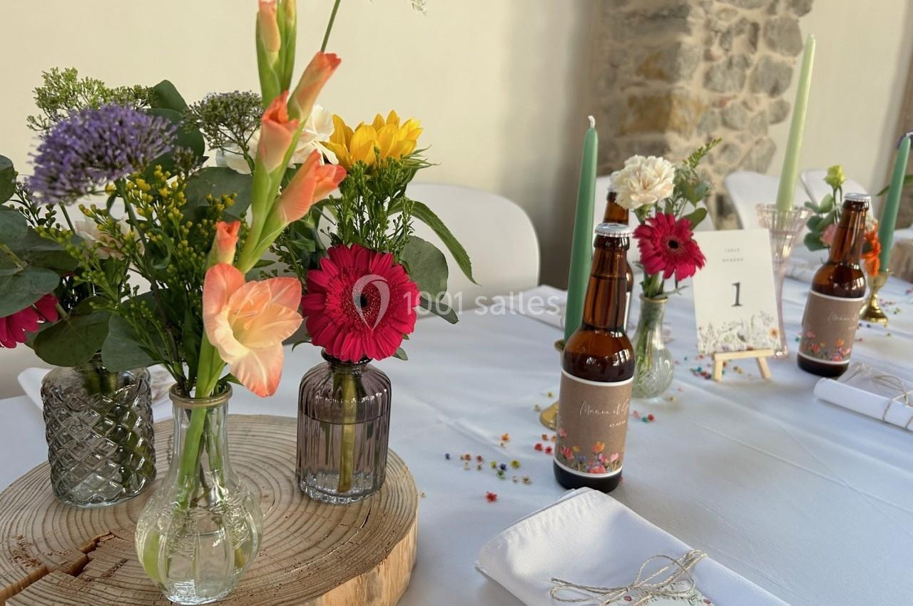 Table décorée avec des fleurs colorées dans des vases, des bougies vertes et des bouteilles en verre sur une nappe blanche.