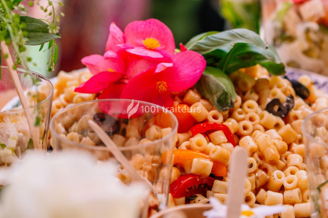 Salade de pâtes garnie de tomates, olives noires et fleurs décoratives roses, entourée de verres et d'herbes fraîches.