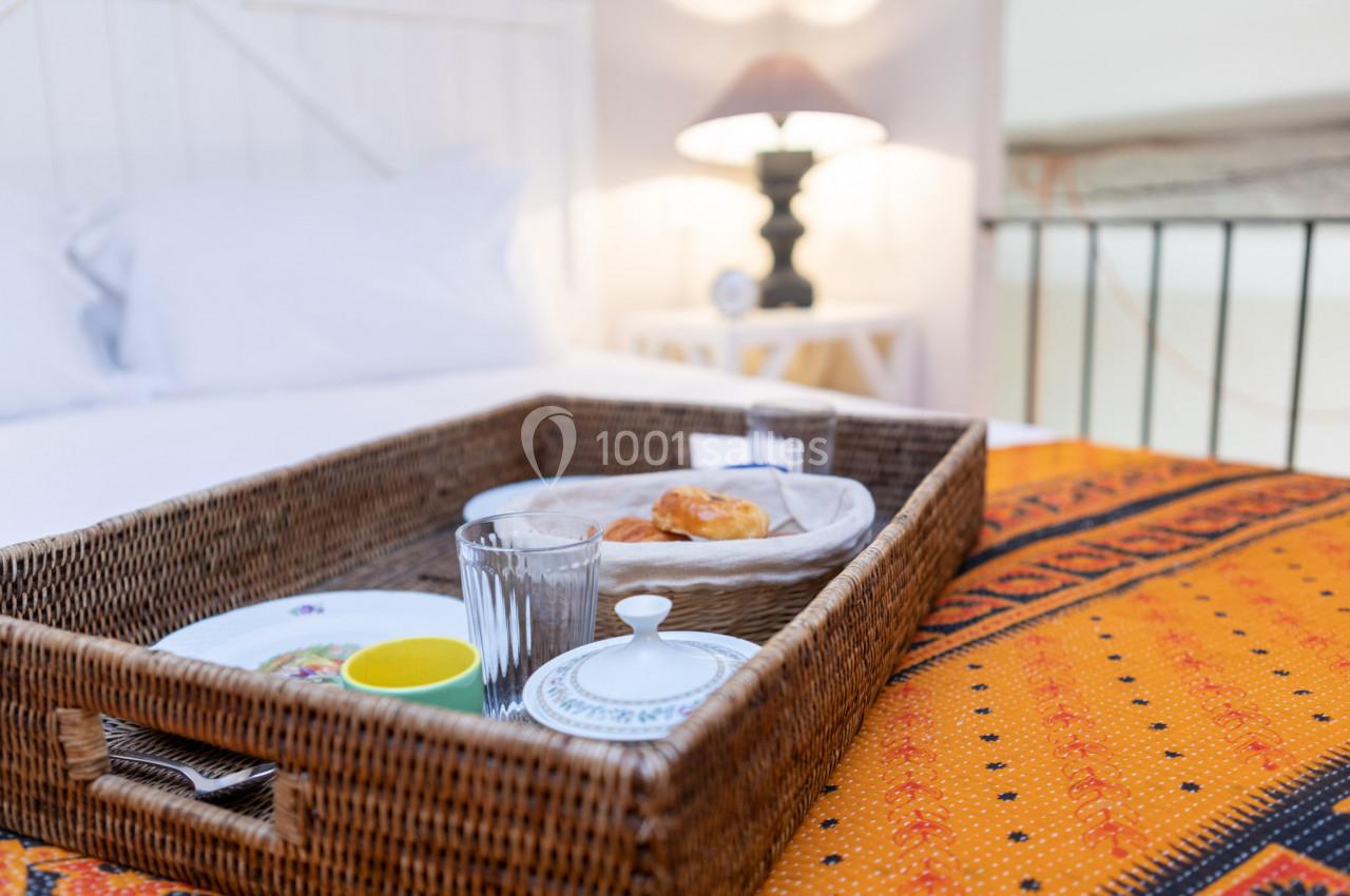 Plateau en osier avec petit-déjeuner, comprenant croissant, vaisselle et verres, posé sur un lit avec couvre-lit orange.