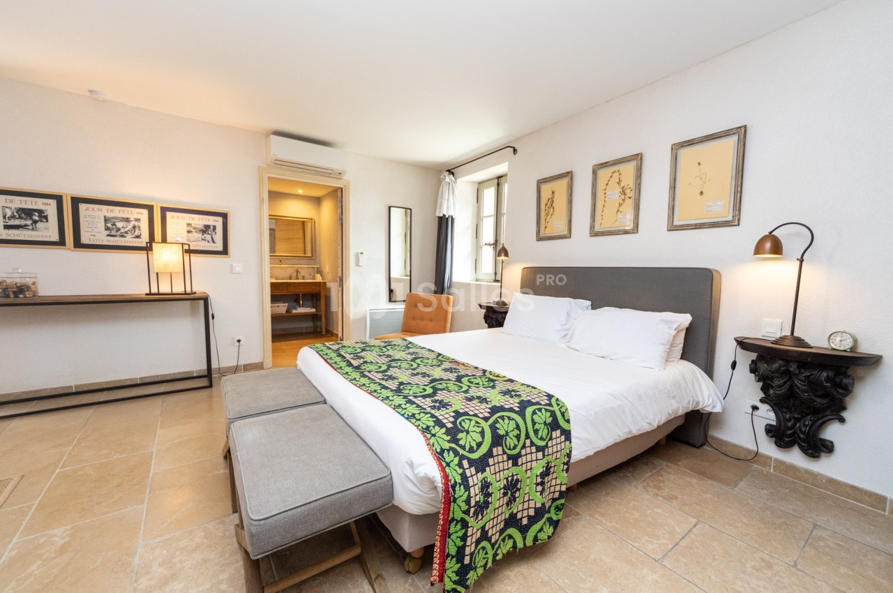Chambre lumineuse avec lit double, banc au pied, décoration murale et salle de bain attenante visible en arrière-plan.