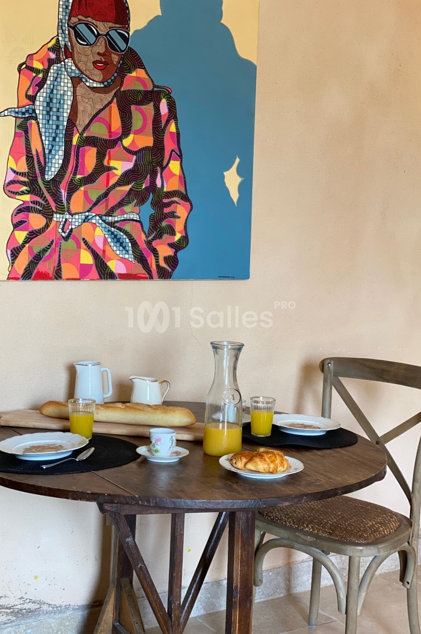 Table en bois avec petit-déjeuner composé de croissants, jus d'orange et café, devant un tableau coloré sur un mur beige.
