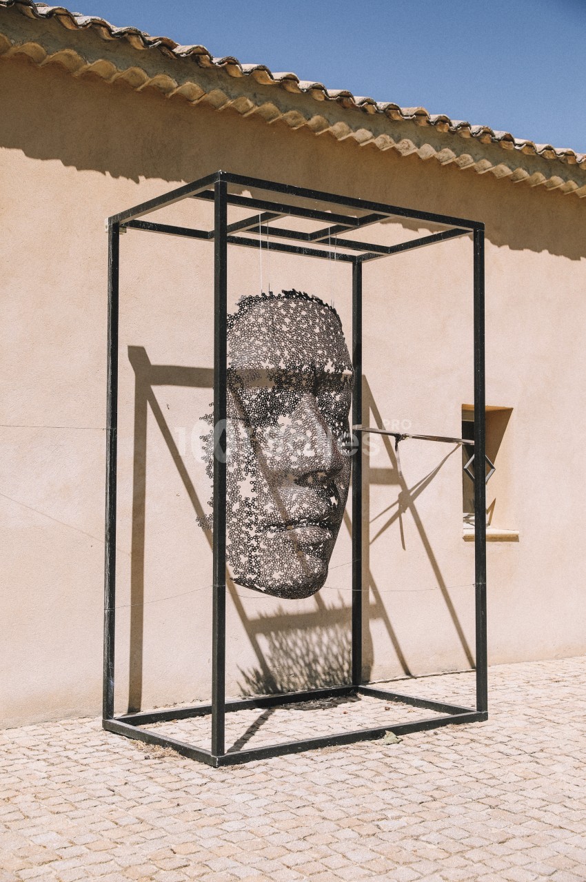 Sculpture suspendue représentant un visage humain, encadrée par une structure métallique, sur une place pavée.