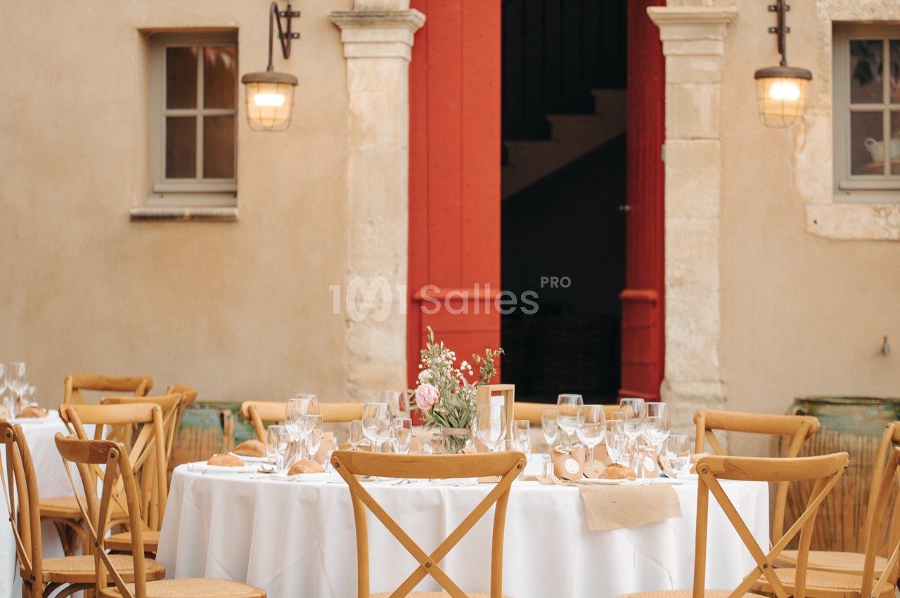 Table ronde dressée avec vaisselle élégante, entourée de chaises en bois, devant une porte rouge et des murs beige.